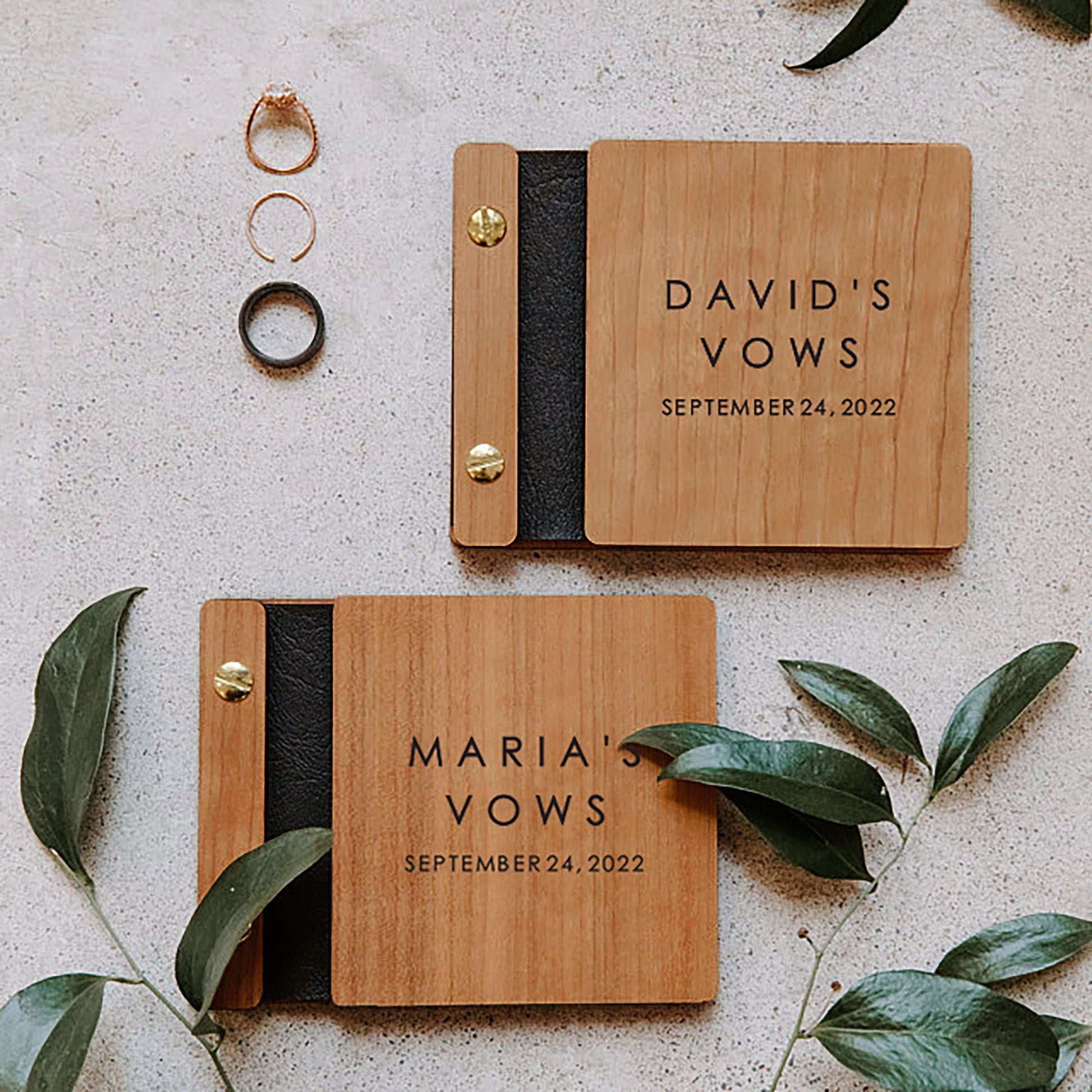 Two vow books made of cherry wood, black vegan leather binding, and gold hardware lie diagonally from one another. Green leaves and golden rings lie next to the vow books as they set the tone of the photo.