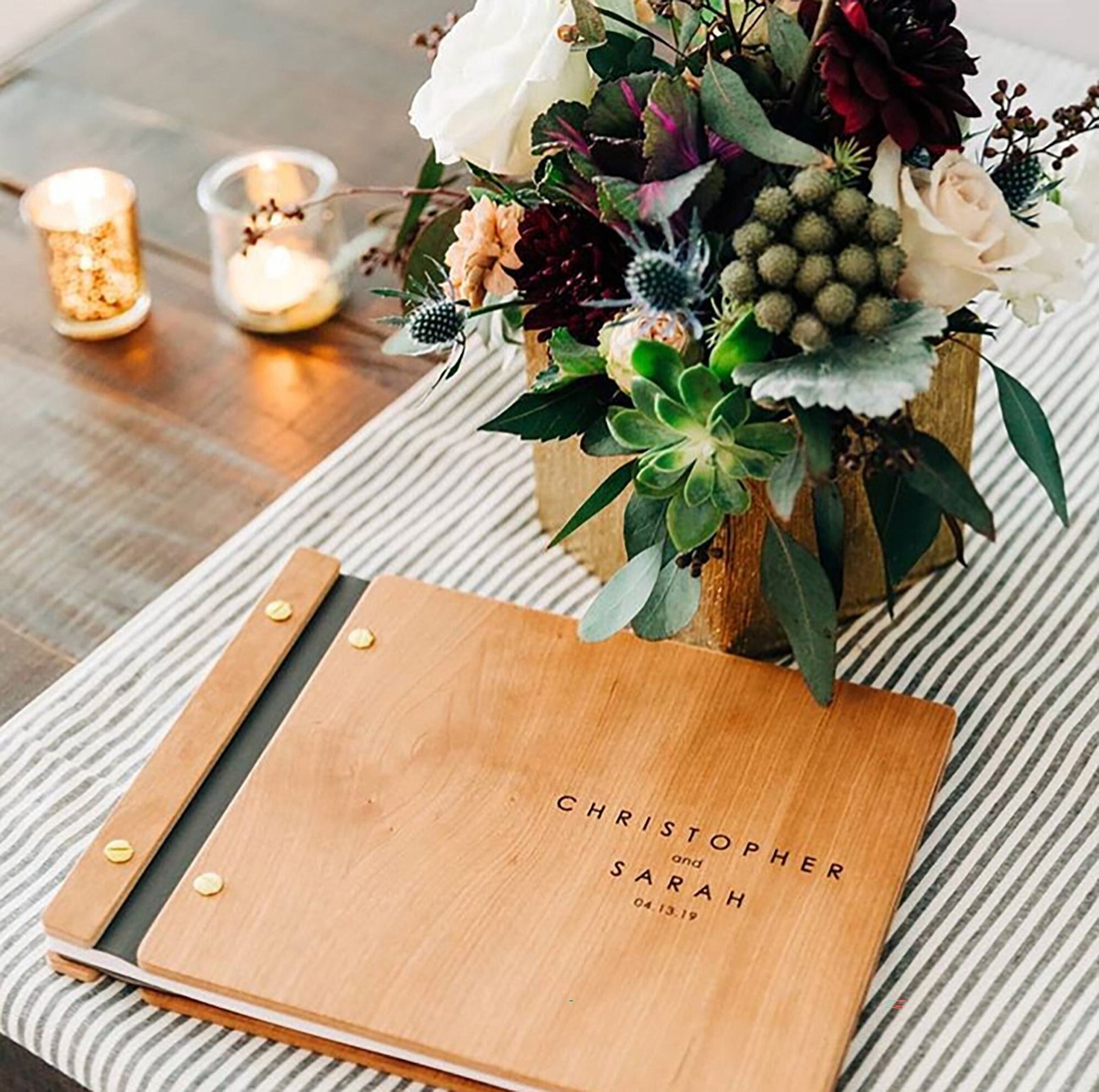 An 8.5x11 inch guest book sits on a striped tablecloth with clear candles and a beautiful bouquet of flowers in the background. It is made of cherry wood, grey vegan leather, and gold screws and reads Christopher and Sarah.