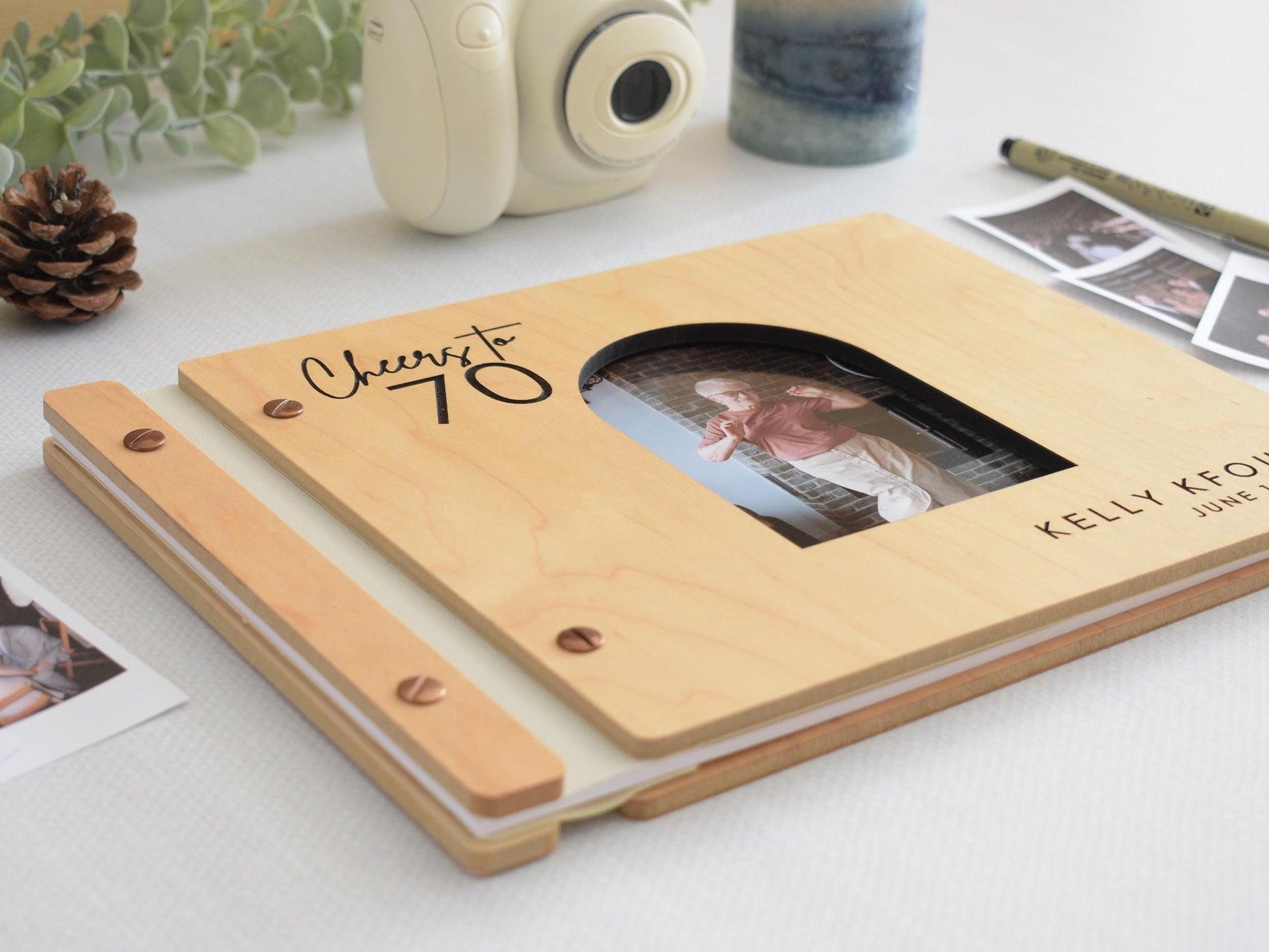 Custom wooden guest book for a 70th birthday celebration. The cover features an engraved &quot;Cheers to 70&quot; message. The book is surrounded by Polaroid pictures, a camera, and decorative elements. 30th 40th 50th 60th 70th 80th 90th birthday