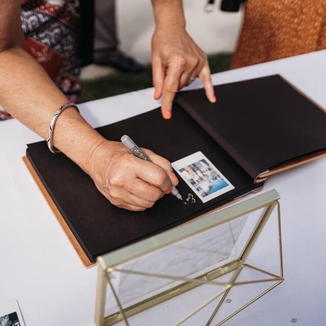 An 8.5x11 inch guest book lies open on a wooden desk. Black cardstock pages with handwritten messages and photo booth strips are posted on the pages for the bride and groom to cherish as a keepsake after their wedding day.