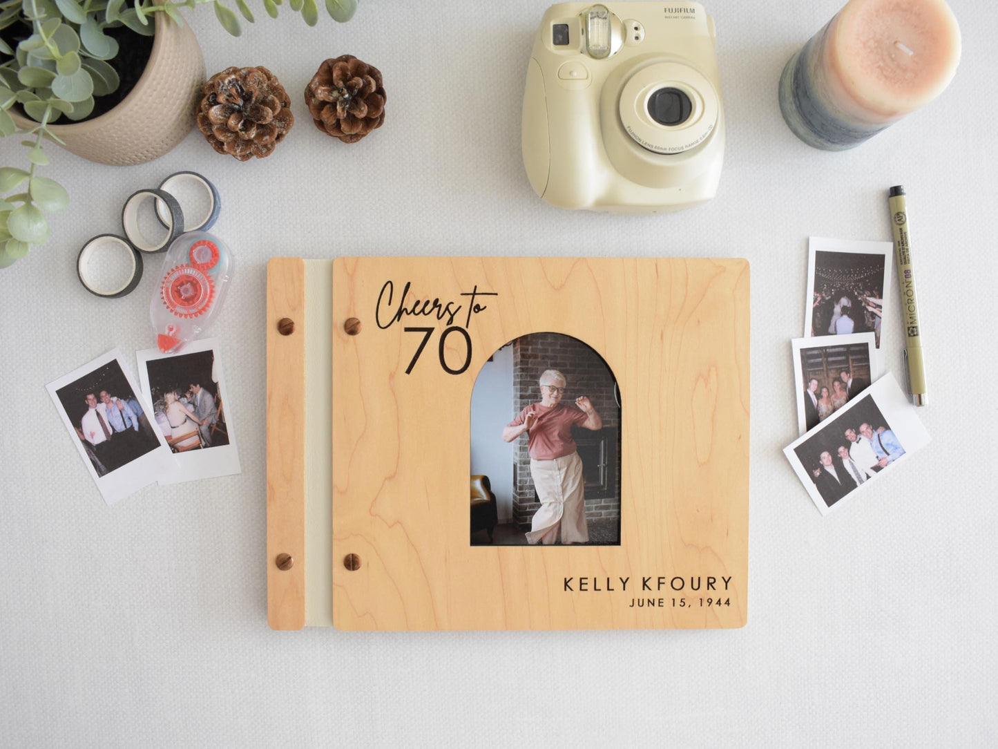 Custom wooden guest book for a 70th birthday celebration. The cover features an engraved &quot;Cheers to 70&quot; message. The book is surrounded by Polaroid pictures, a camera, and decorative elements. 30th 40th 50th 60th 70th 80th 90th birthday