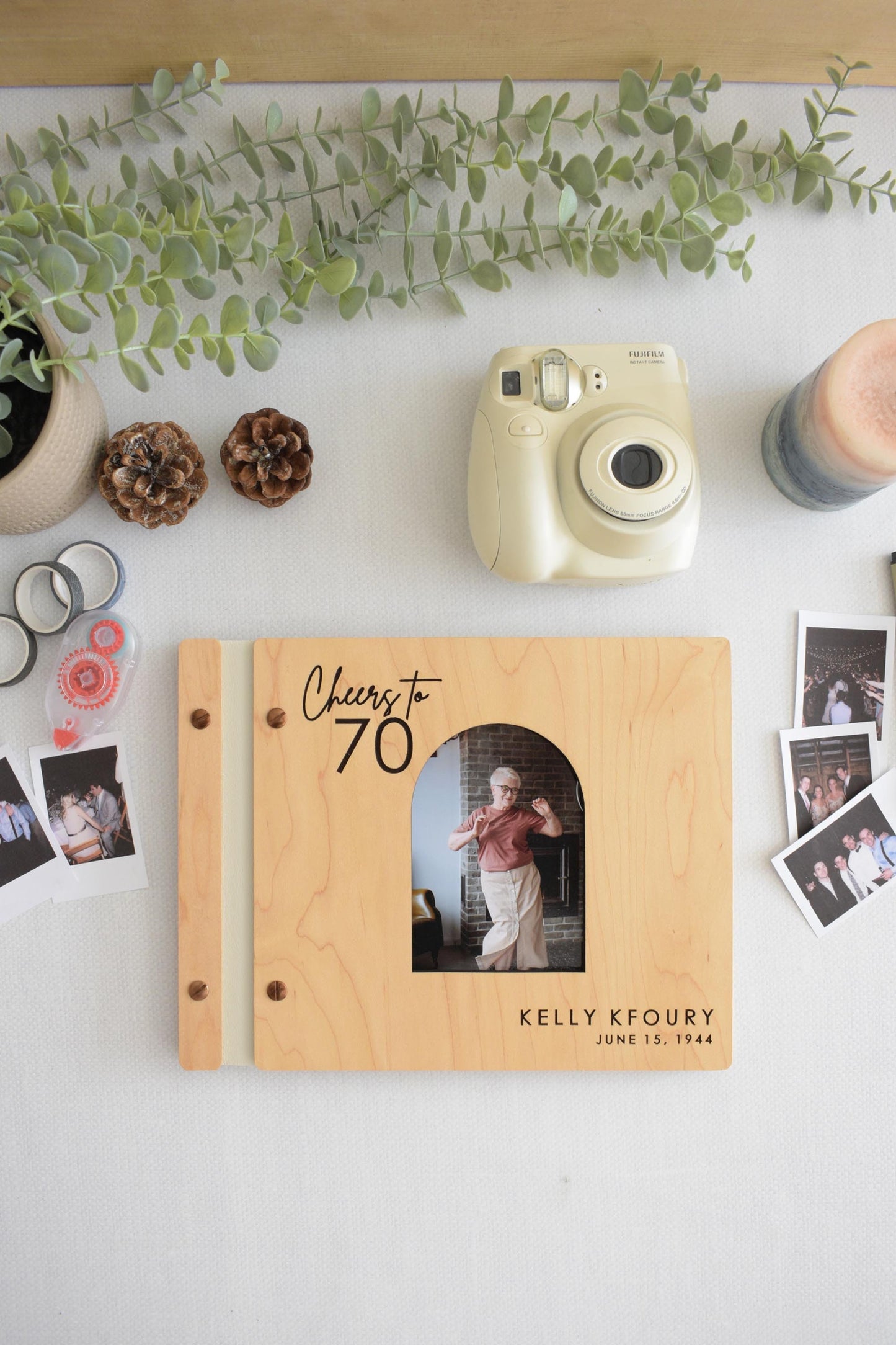 Custom wooden guest book for a 70th birthday celebration. The cover features an engraved &quot;Cheers to 70&quot; message. The book is surrounded by Polaroid pictures, a camera, and decorative elements. 30th 40th 50th 60th 70th 80th 90th birthday